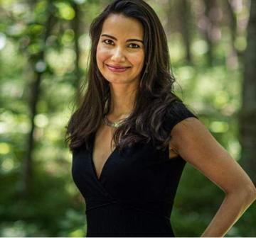 woman standing side on in black dress with long brown hair smiles at the camera. Green blurred background