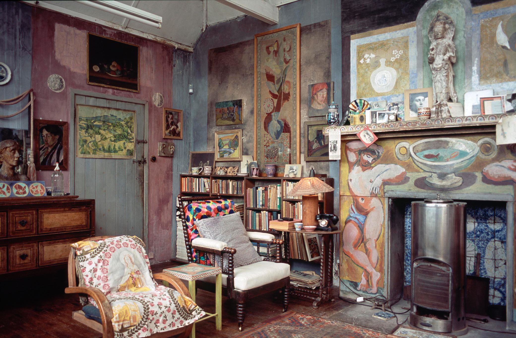 Charleston A cluttered corner of an indoor art studio - walls painted in various block colours, two chairs with multicoloured cushions, paintings and sculptures covering the walls. A bookshelf in the background and an unlit fireplace with a tiled and painted facade
